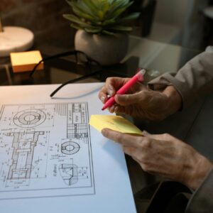 A person annotating a technical blueprint in a modern workspace setting with a laptop and coffee.